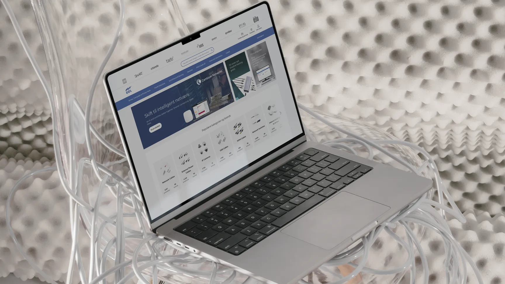 A MacBook laptop displaying a website on intelligent networking, surrounded by tangled cables, resting in a transparent chair with a textured background.