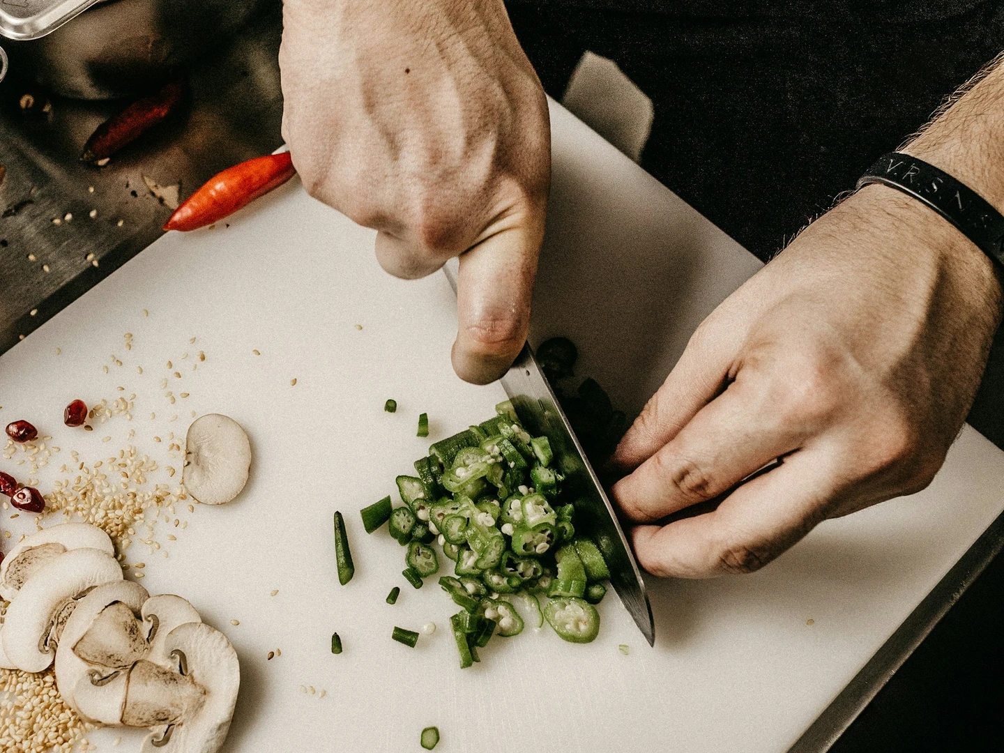 Chef chopping green onions on a cutting board, surrounded by fresh ingredients like salmon, mushrooms, and sesame seeds in a professional kitchen setting.