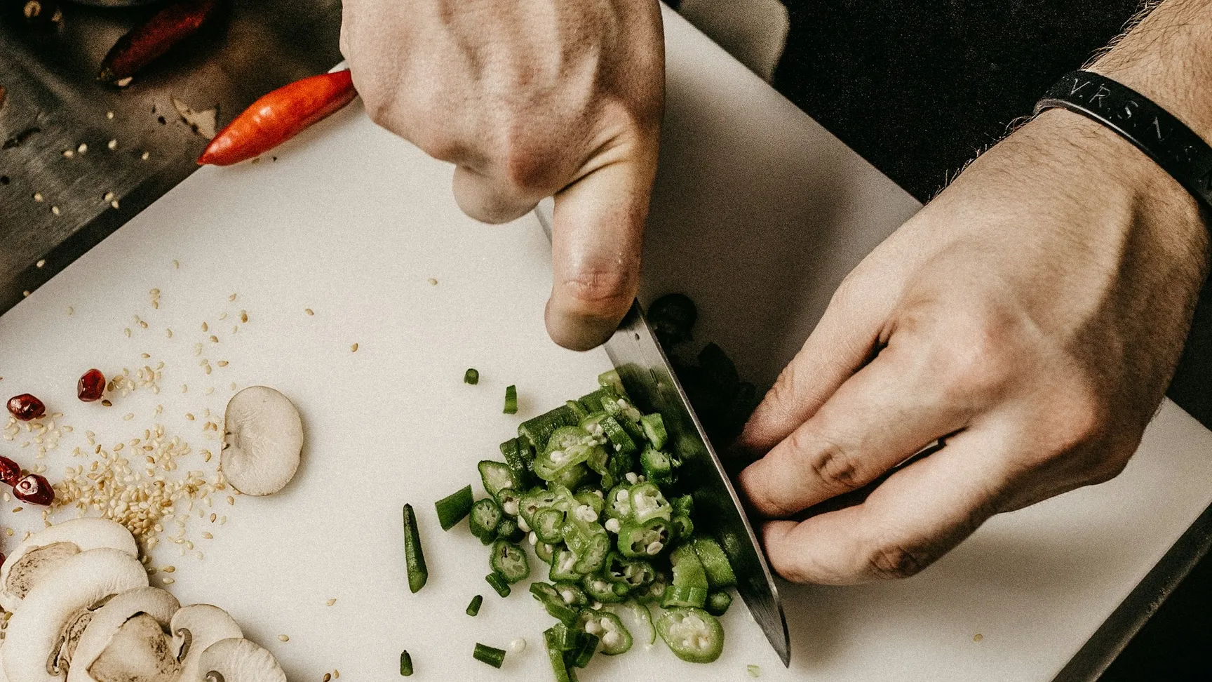 Chef chopping green onions on a cutting board, surrounded by fresh ingredients like salmon, mushrooms, and sesame seeds in a professional kitchen setting.