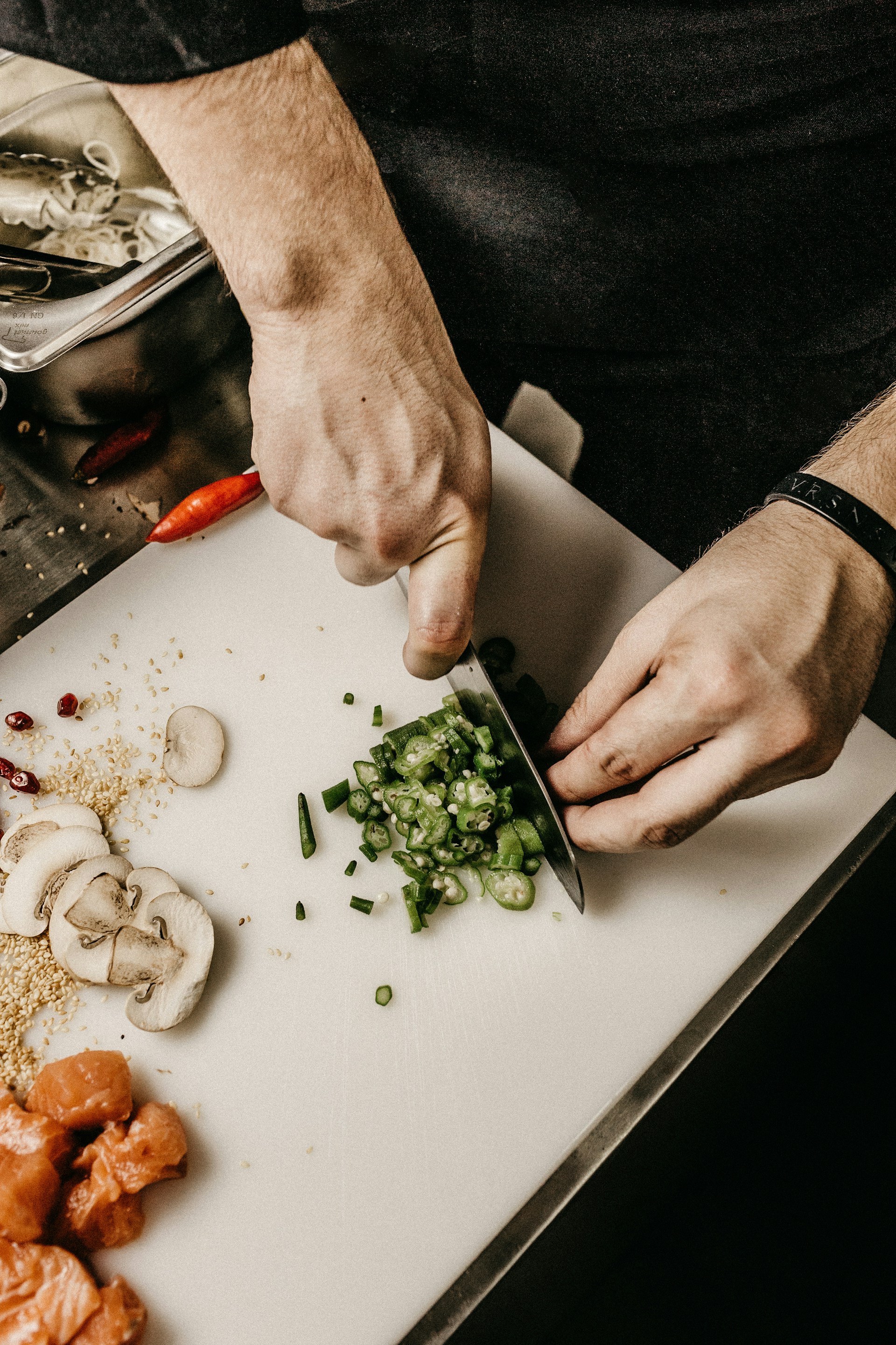 En kok skærer grønne løg på et skærebræt i et moderne køkken, omringet af forskellige ingredienser som svampe, sesamfrø og røde chili.