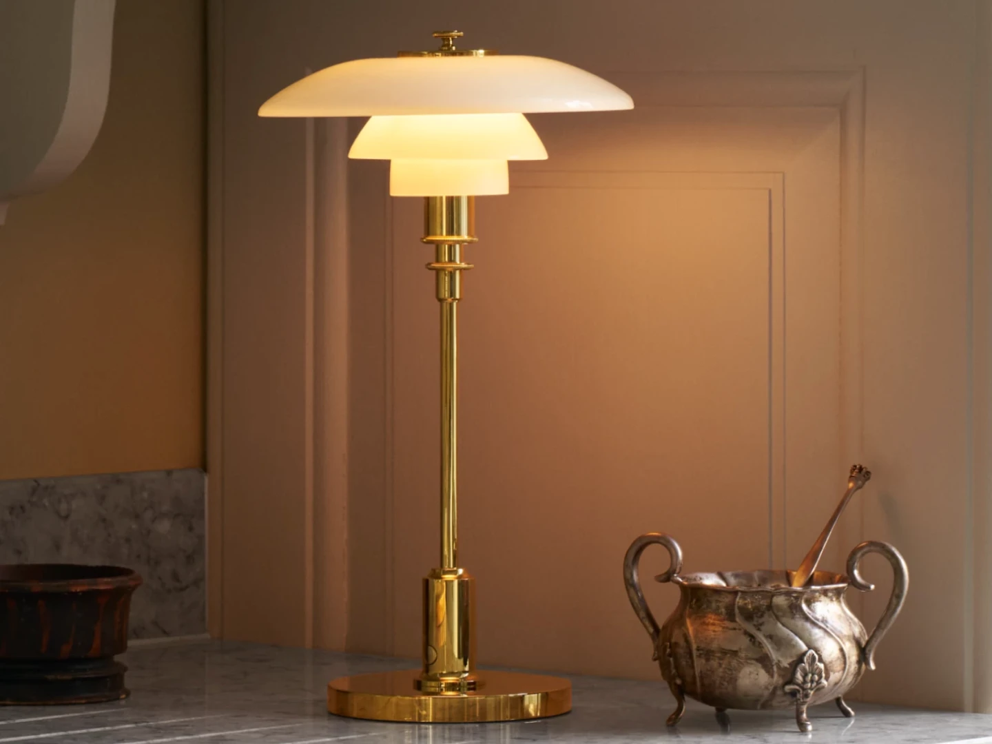 A stylish brass table lamp with a layered white glass shade, positioned on a marble countertop next to an ornate silver bowl and a wooden utensil, creating an elegant kitchen vignette.