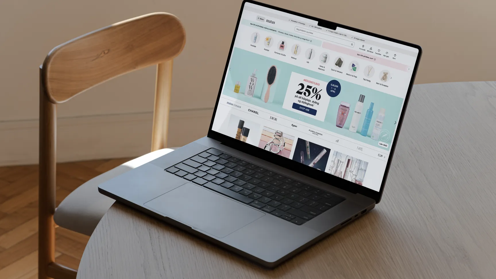 A modern laptop displaying a beauty product website with a promotional banner for 25% off, placed on a wooden table next to a simple chair.