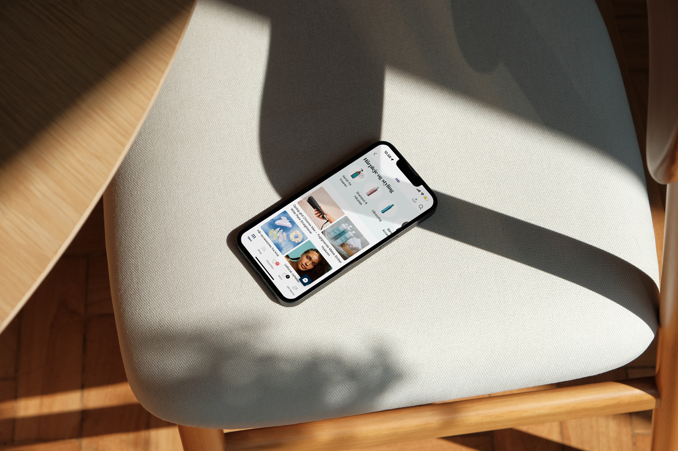 Smartphone der ligger på en lys stol, viser en app med billeder af skønhedsprodukter og kvinder, om sommer. Sollys kaster skygger på stolen.