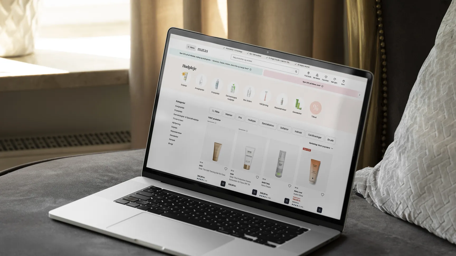 A laptop displaying a skincare product webpage on a soft gray sofa with natural light coming through a window, emphasizing online shopping for beauty products.