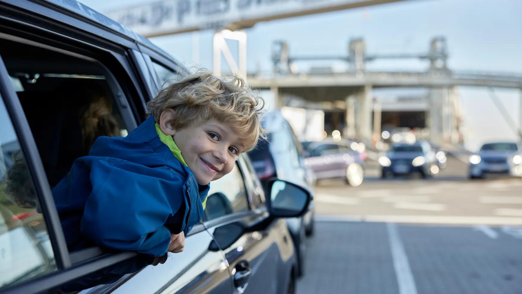 Dreng har hovdet ude af vinduet på en bil, bilen holder bag en anden bil ved en moderne havn