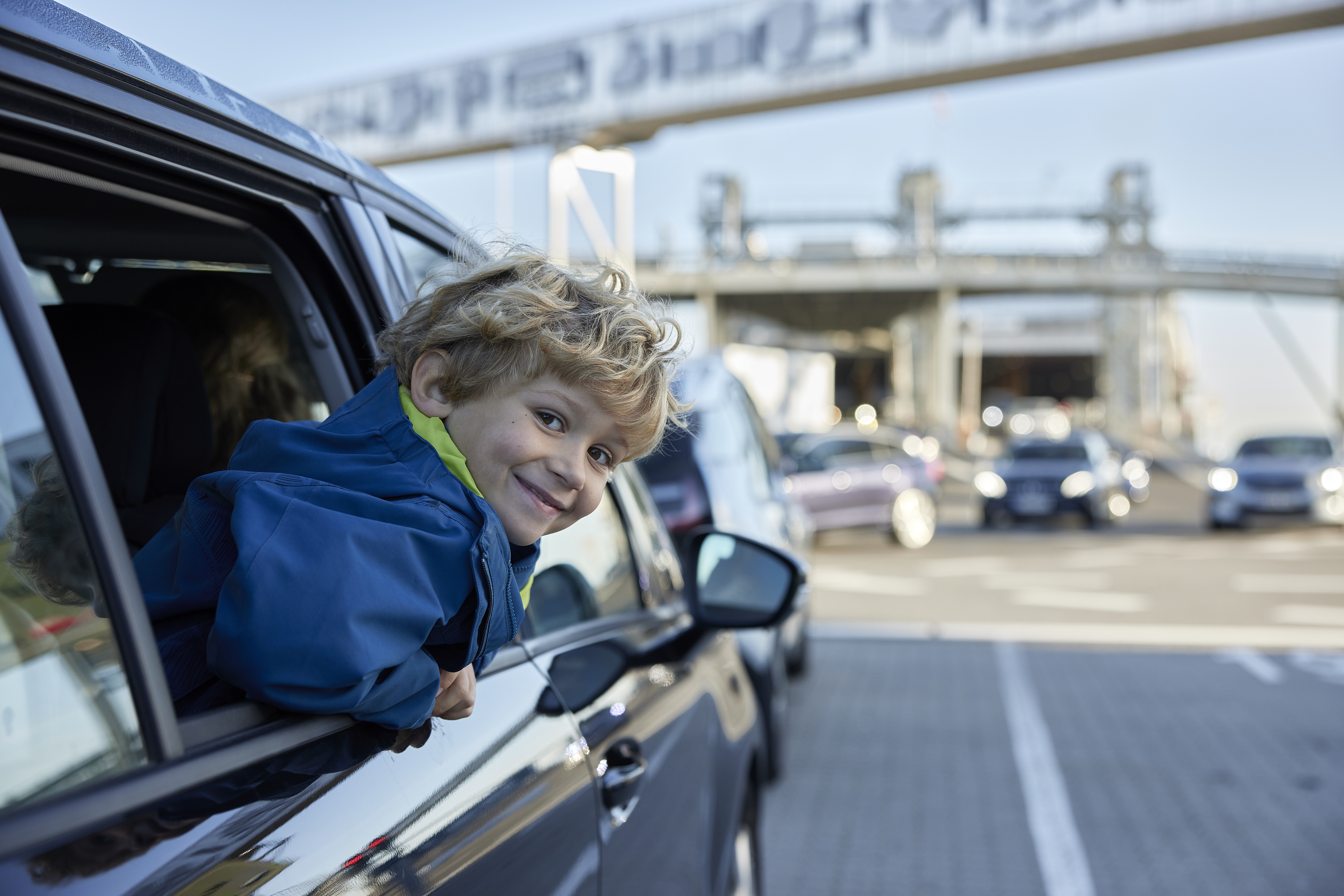 Dreng har hovdet ude af vinduet på en bil, bilen holder bag en anden bil ved en moderne havn