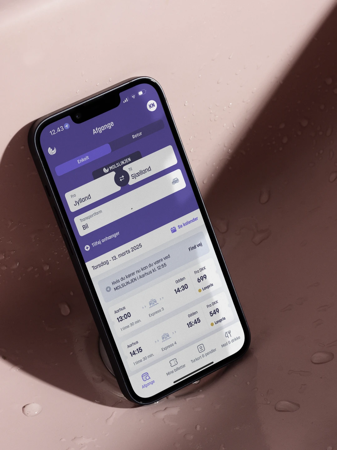 Billede af en smartphone, der viser en rejseapp med information om afgange fra Jylland til Sjælland, placeret i en pink håndvask med dråber på overfladen.