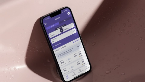 Billede af en smartphone, der viser en rejseapp med information om afgange fra Jylland til Sjælland, placeret i en pink håndvask med dråber på overfladen.