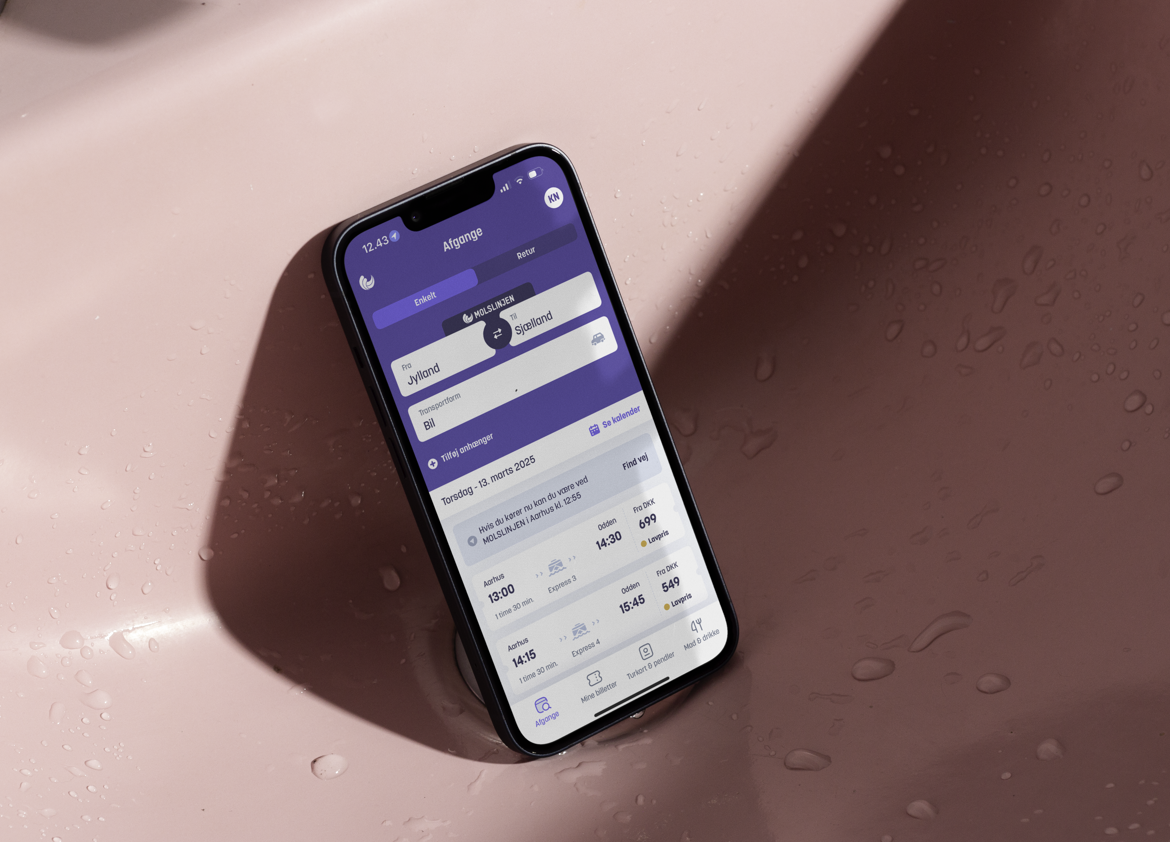 Billede af en smartphone, der viser en rejseapp med information om afgange fra Jylland til Sjælland, placeret i en pink håndvask med dråber på overfladen.