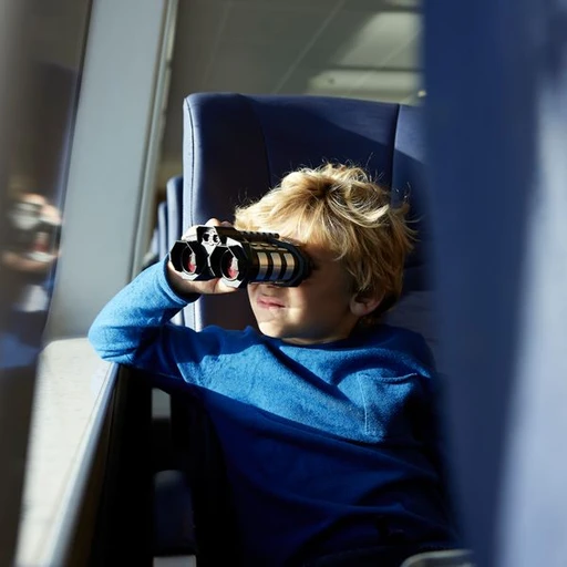 A young boy with tousled blond hair is sitting by a window, holding binoculars up to his eyes, engaging in exploration and observation, wearing a blue long-sleeve shirt.