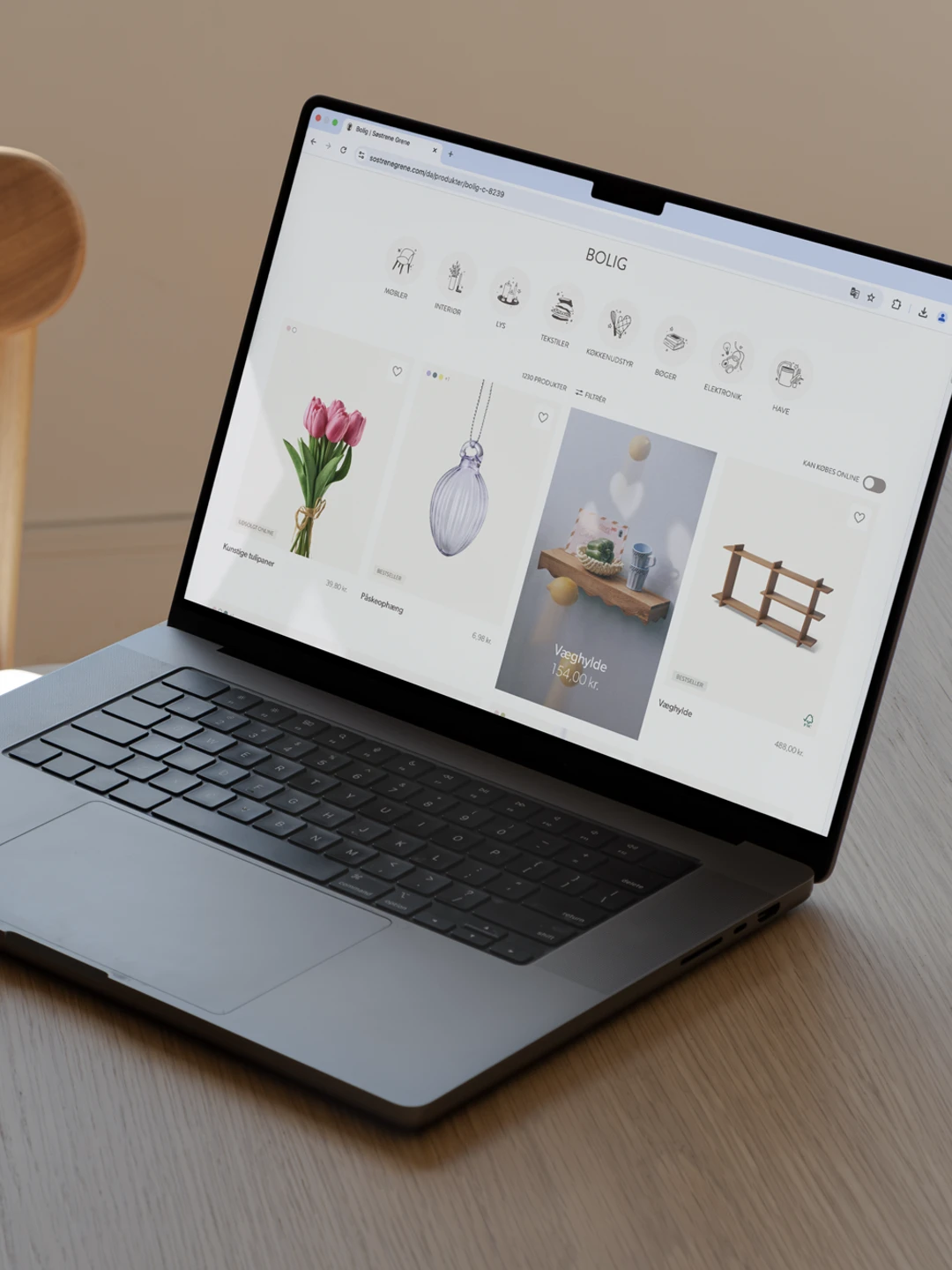 Billede af en bærbar computer, der viser en boligindretning hjemmeside med blomster og dekorative genstande, placeret på et rundt træbord med en stol i baggrunden.
