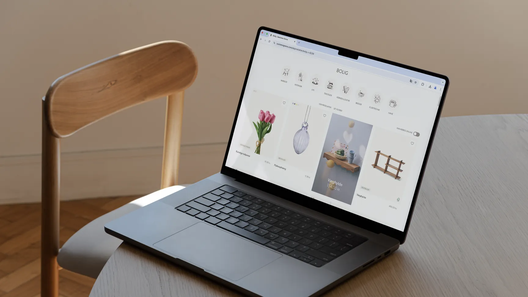 Billede af en bærbar computer, der viser en boligindretning hjemmeside med blomster og dekorative genstande, placeret på et rundt træbord med en stol i baggrunden.