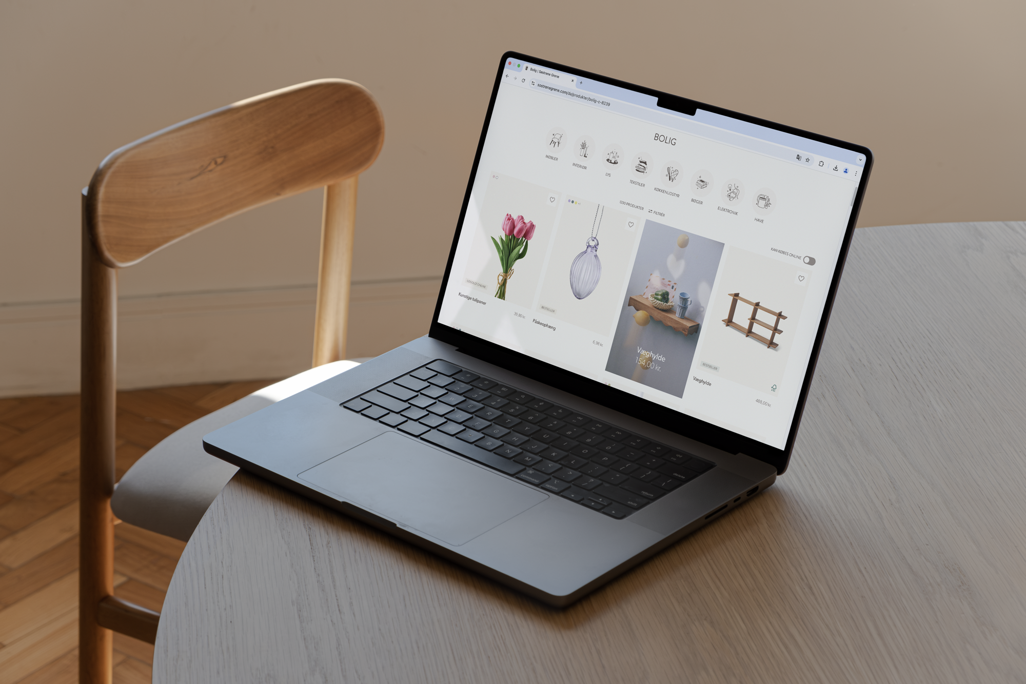 Billede af en bærbar computer, der viser en boligindretning hjemmeside med blomster og dekorative genstande, placeret på et rundt træbord med en stol i baggrunden.