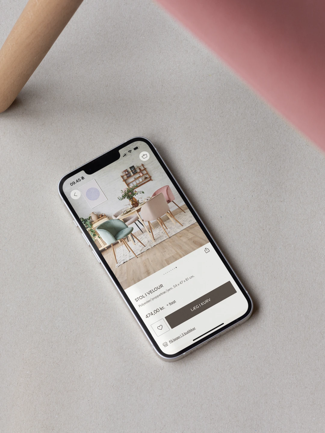 En smartphone liggende på en lys overflade, viser en app med billeder af farverige stole, mens en del af en stol i rosa vises i billedets øverste højre hjørne.