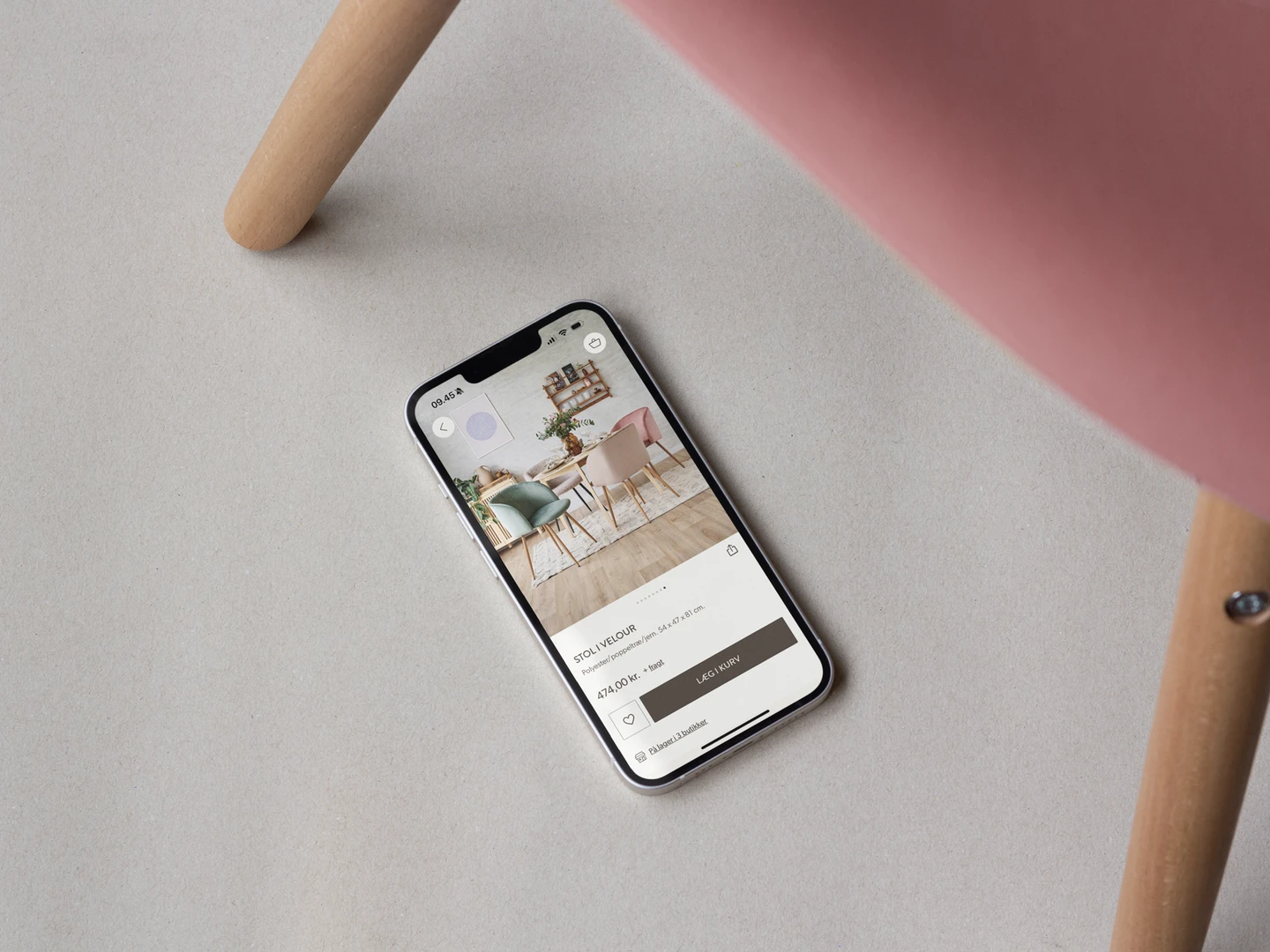 En smartphone liggende på en lys overflade, viser en app med billeder af farverige stole, mens en del af en stol i rosa vises i billedets øverste højre hjørne.