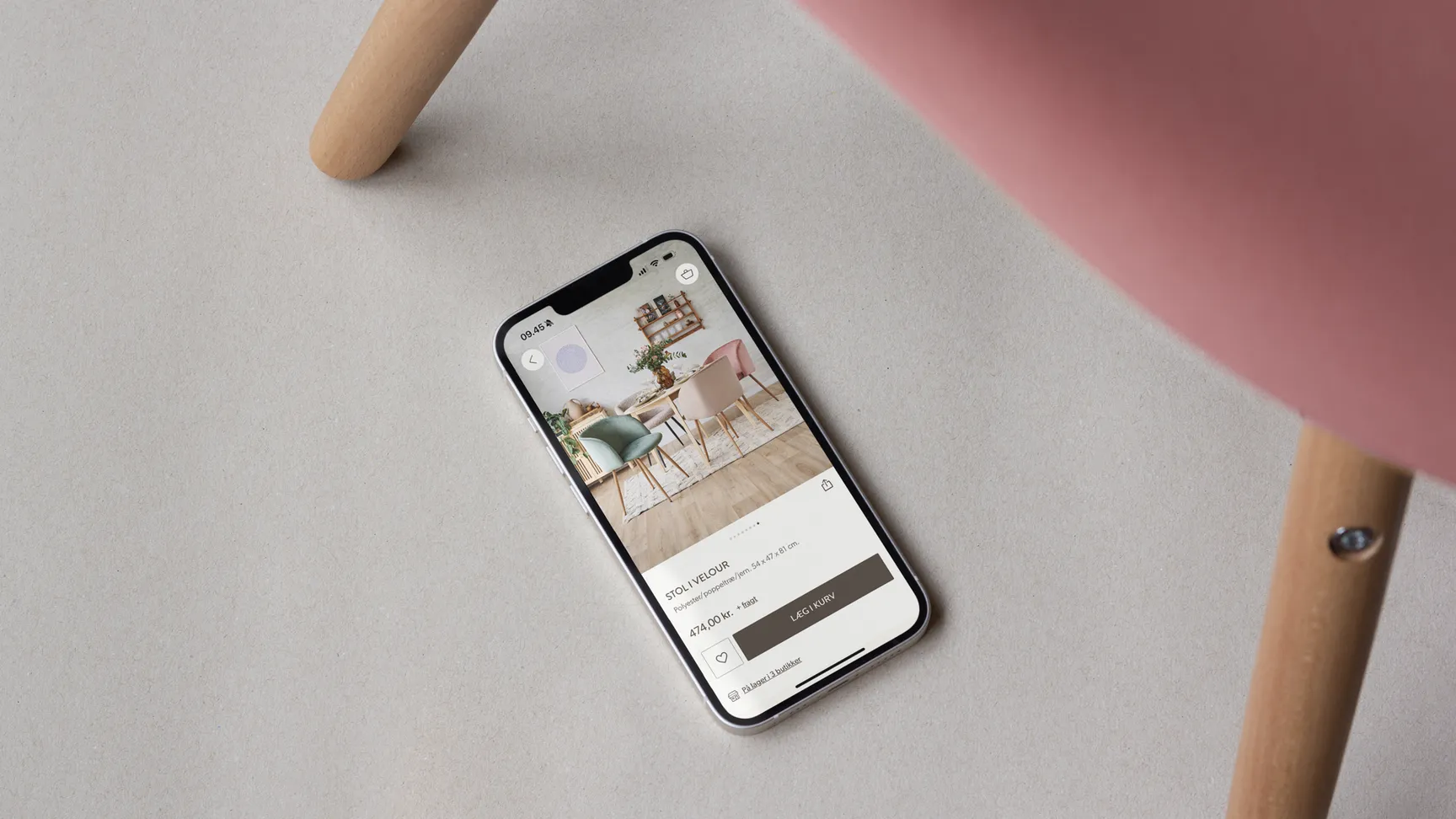 En smartphone liggende på en lys overflade, viser en app med billeder af farverige stole, mens en del af en stol i rosa vises i billedets øverste højre hjørne.