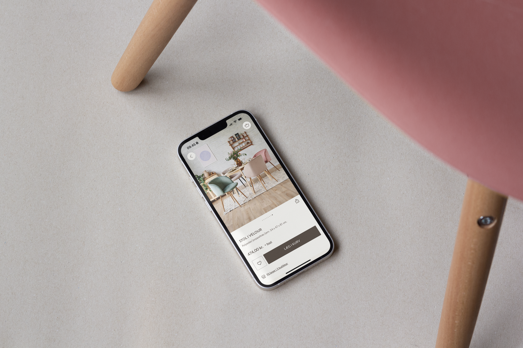 En smartphone liggende på en lys overflade, viser en app med billeder af farverige stole, mens en del af en stol i rosa vises i billedets øverste højre hjørne.