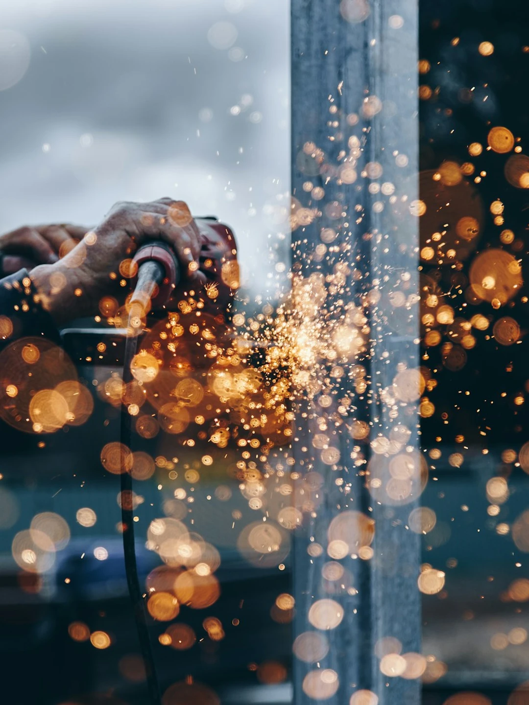 A person using a power tool to create sparks while working on metal, with a blurred background featuring bright bokeh lights and a cloudy sky.