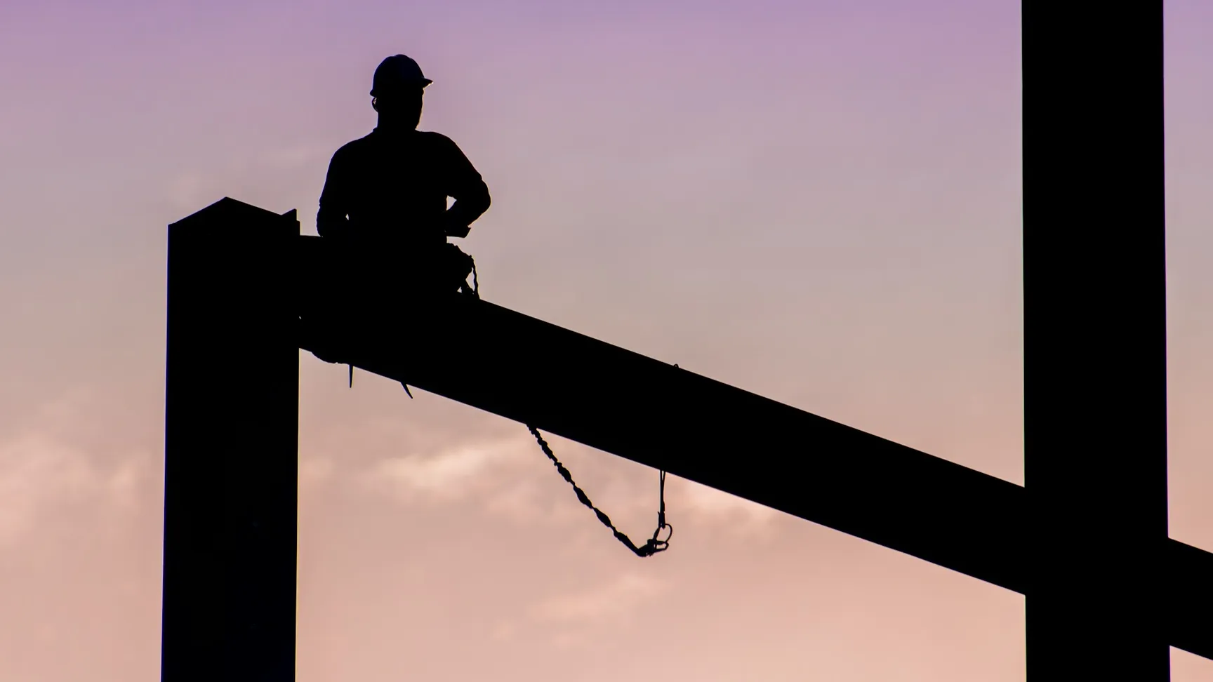 Silhouette of a construction worker sitting on a steel beam against a sunset sky, emphasizing safety in construction.