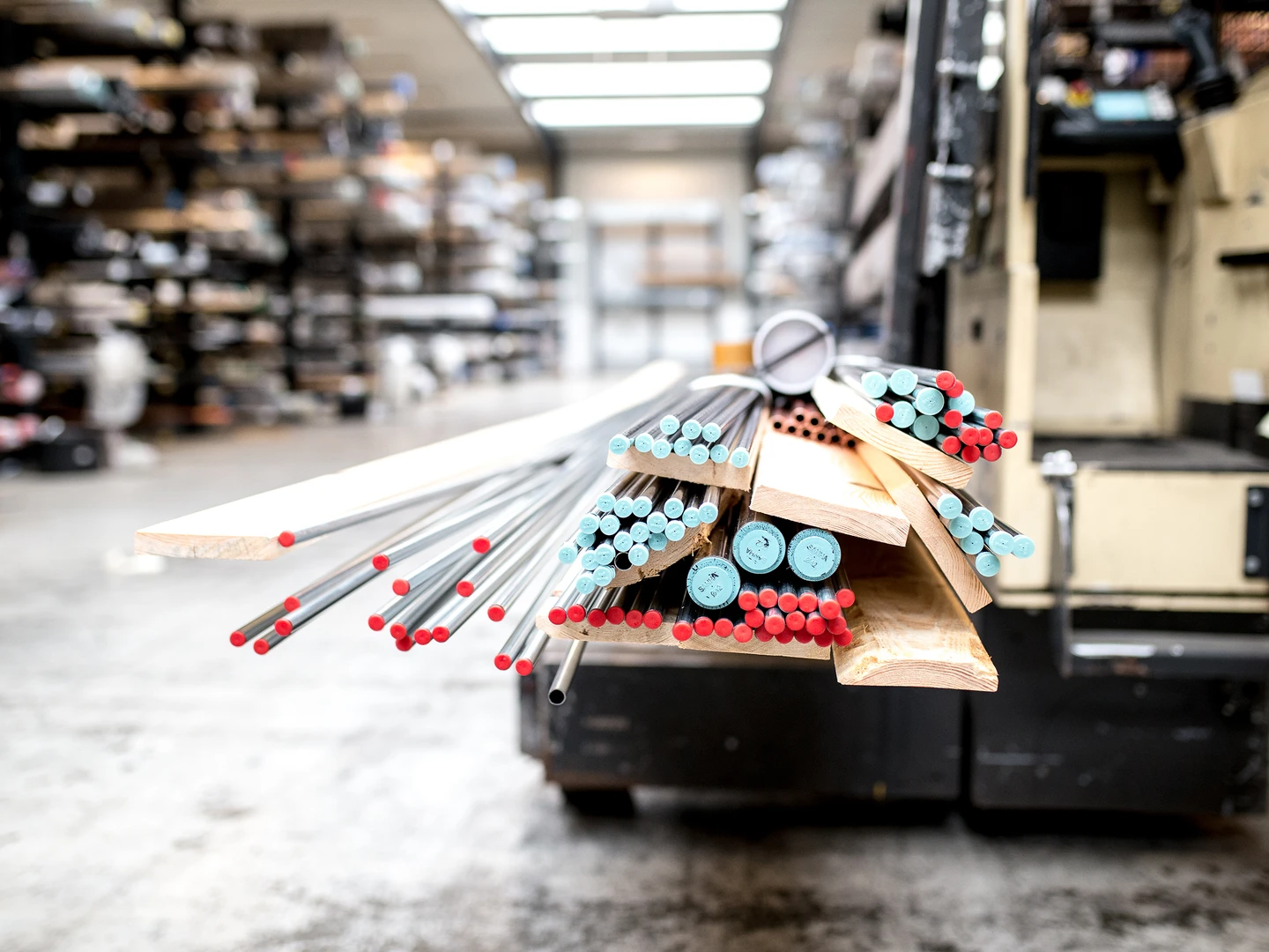 Various metal rods, PVC pipes, and wooden planks stacked on a forklift in a warehouse, with shelves filled with materials in the background.
