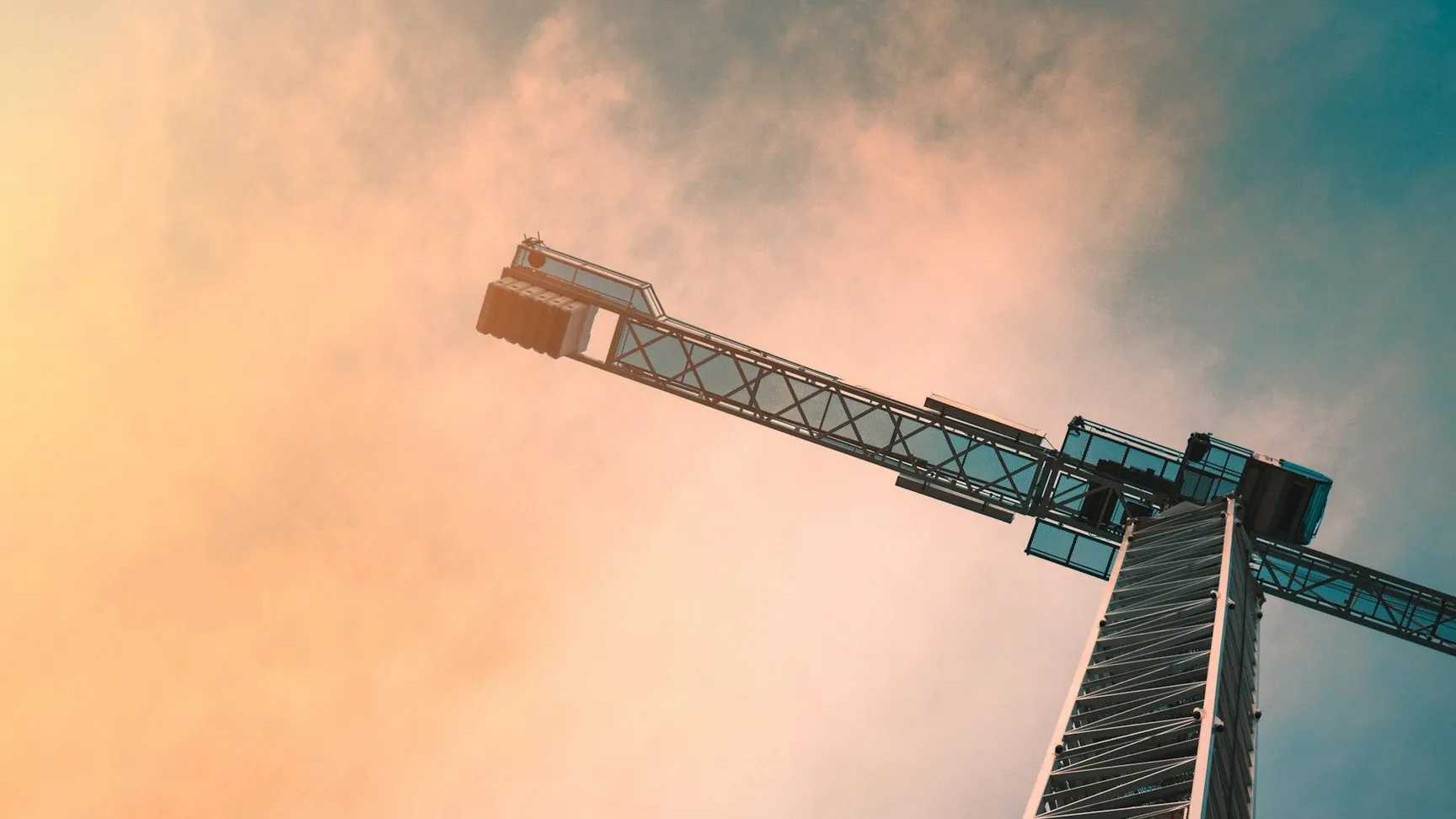 En høj byggekran strækker sig op mod en farverig himmel med skyer, mens den står ved siden af en bygning i konstruktion.