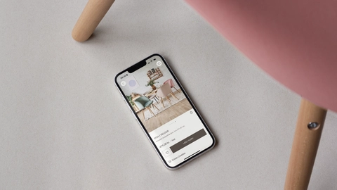 En smartphone liggende på en lys overflade, viser en app med billeder af farverige stole, mens en del af en stol i rosa vises i billedets øverste højre hjørne.
