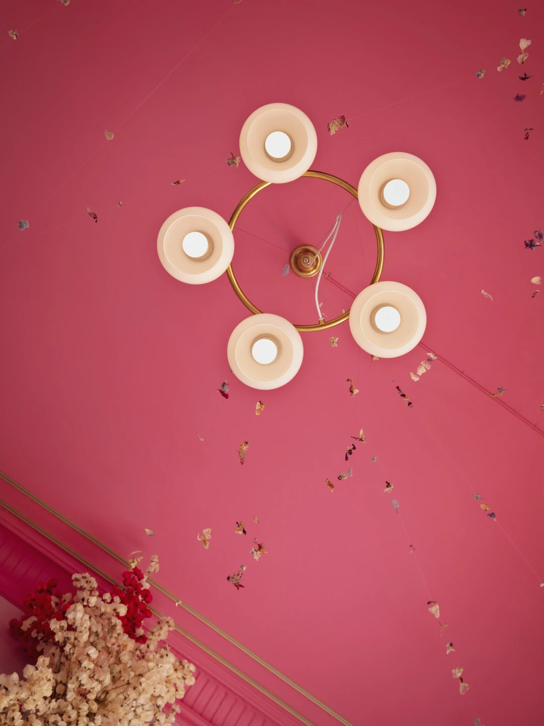 Ceiling view featuring a circular gold light fixture with five white shades against a vibrant pink background, adorned with colorful floral decorations hanging by threads.