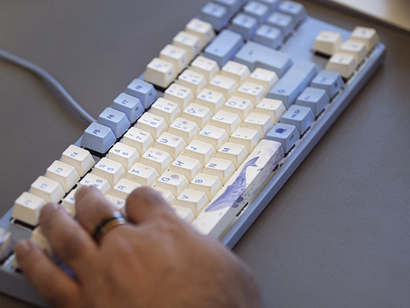 Hånd der skriver på et mekanisk tastatur med blå og hvide taster, med en bærbar computer i baggrunden.