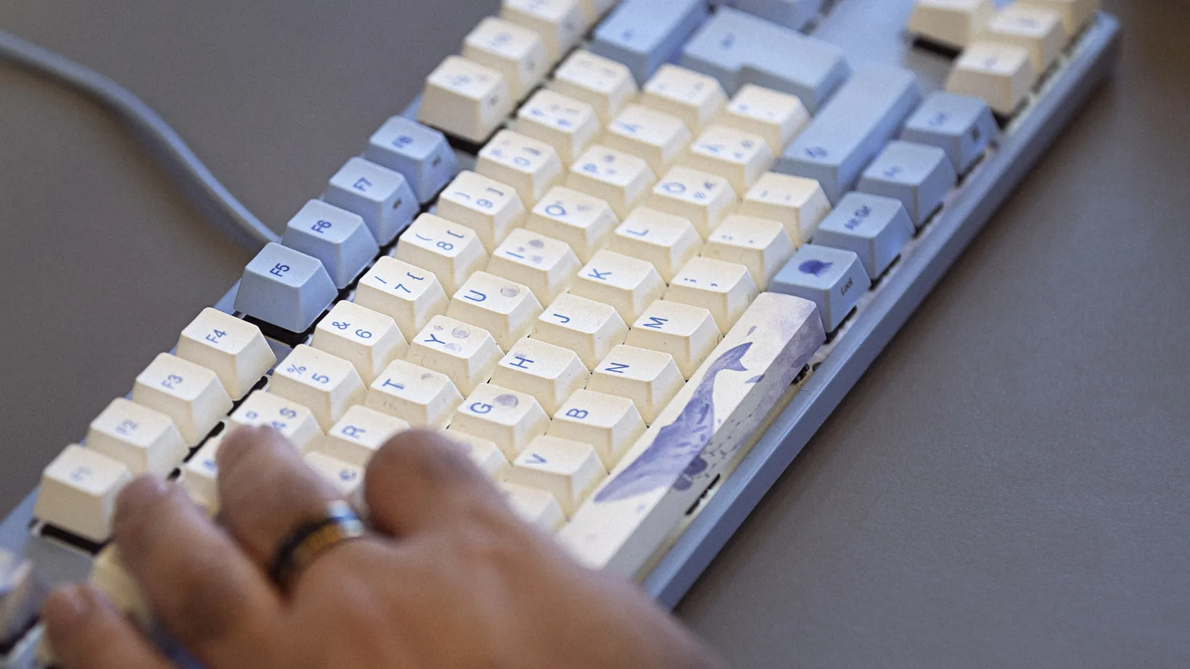 Hånd der skriver på et mekanisk tastatur med blå og hvide taster, med en bærbar computer i baggrunden.