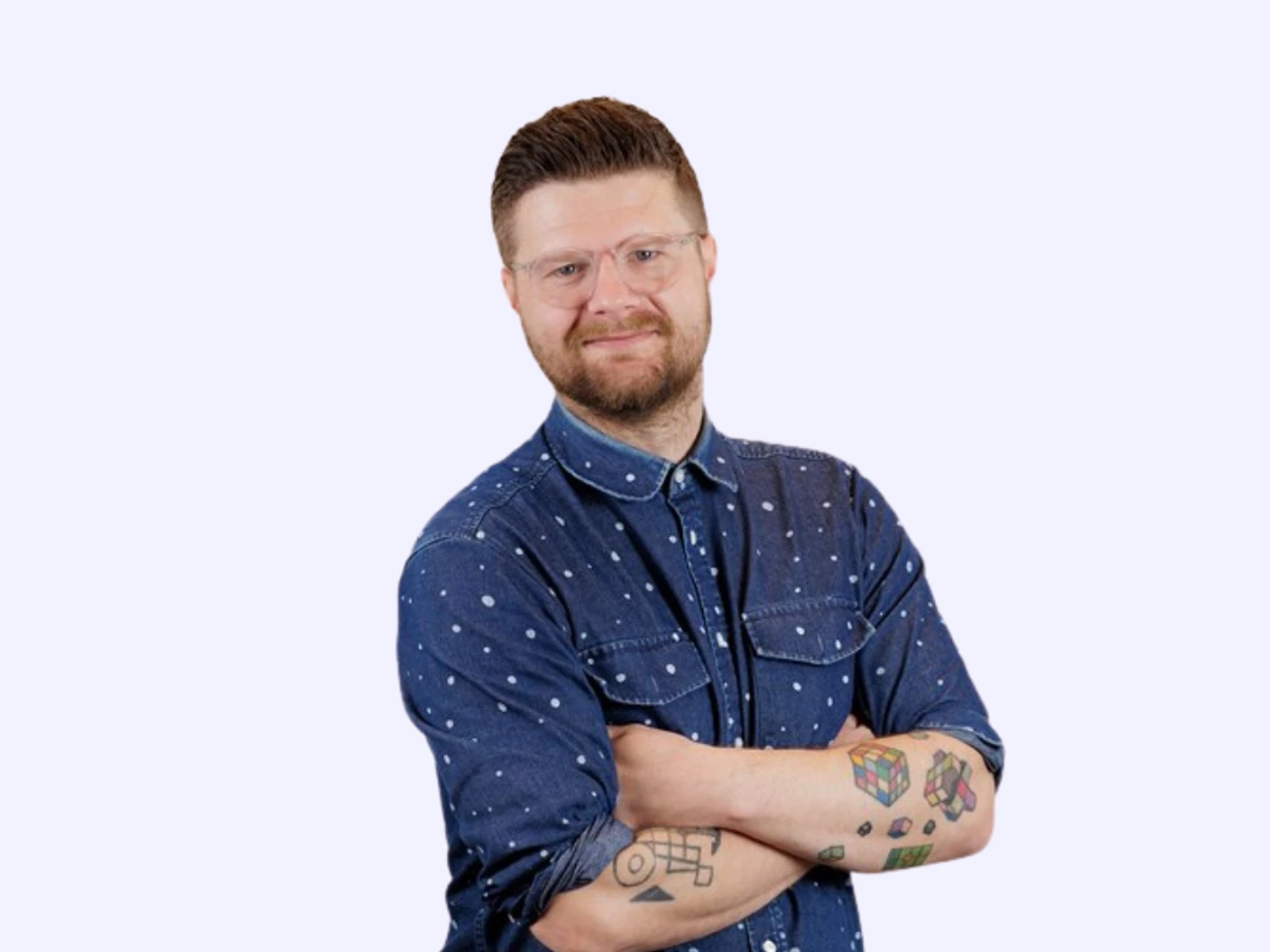 En mand med briller og tatoveringer, iført en denimskjorte med prikker, står med armene korslagt og smiler mod kameraet.