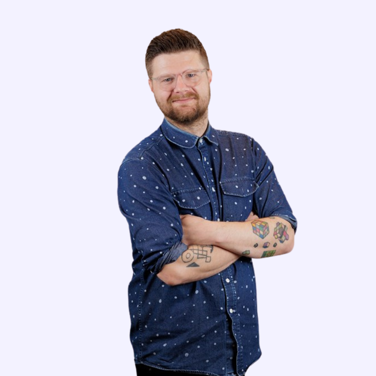 En mand med briller og tatoveringer, iført en denimskjorte med prikker, står med armene korslagt og smiler mod kameraet.