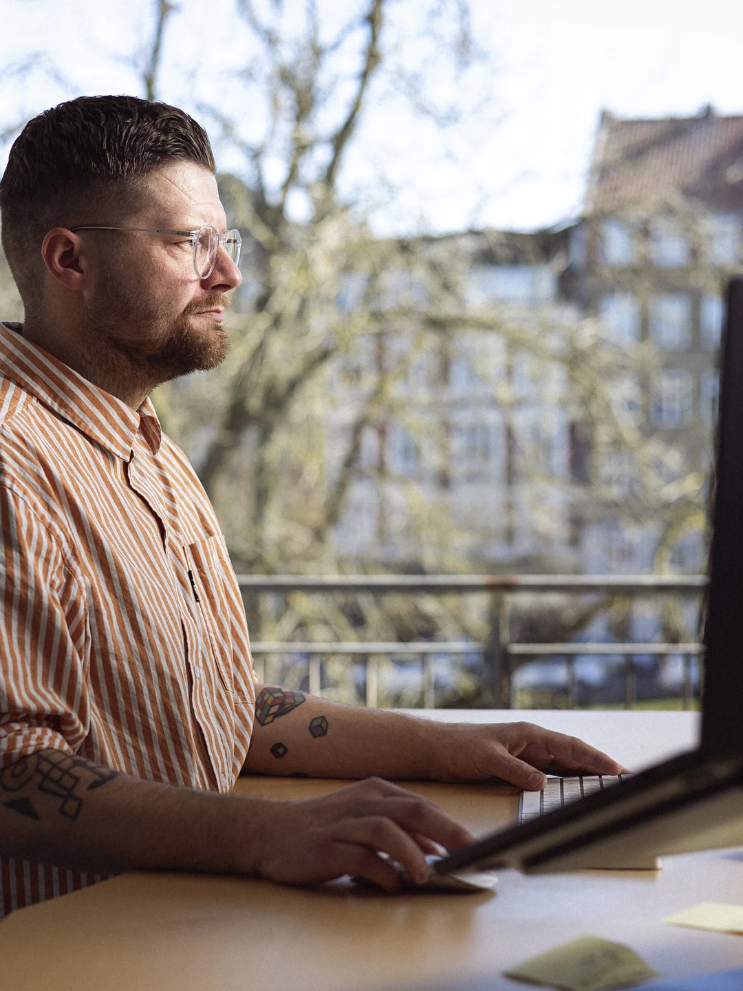 Mand arbejder på en computer ved et skrivebord med en balkonudsigt, iført en stribet skjorte.