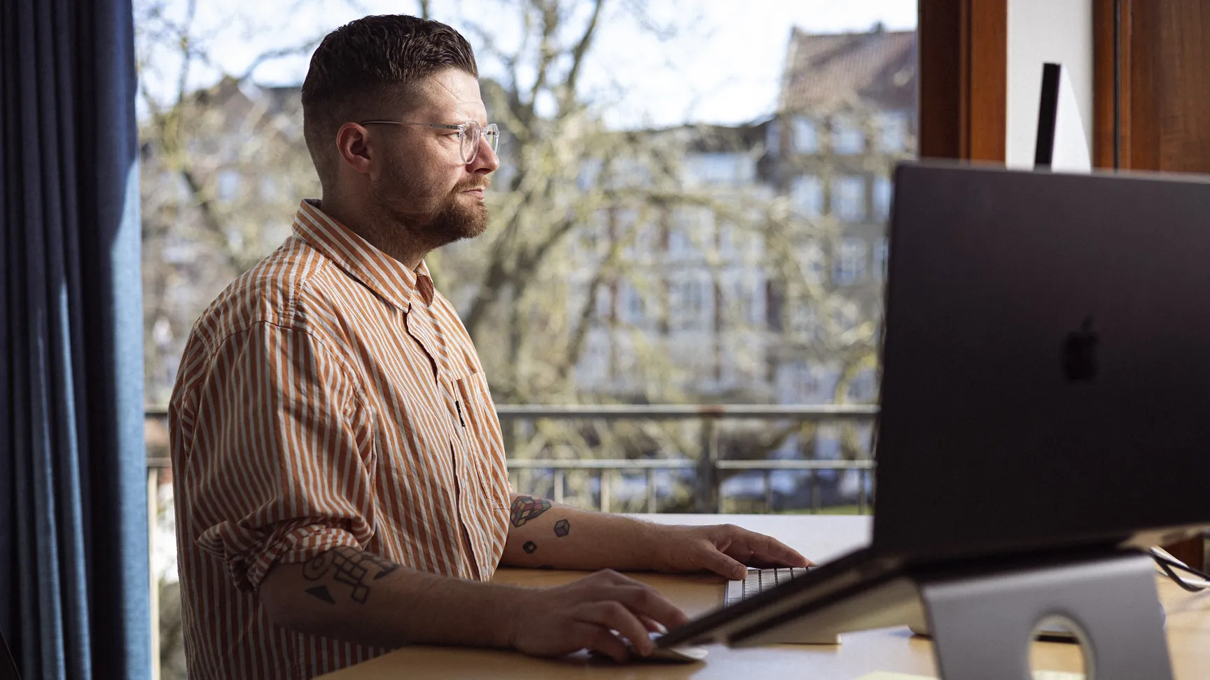 Mand arbejder på en computer ved et skrivebord med en balkonudsigt, iført en stribet skjorte.