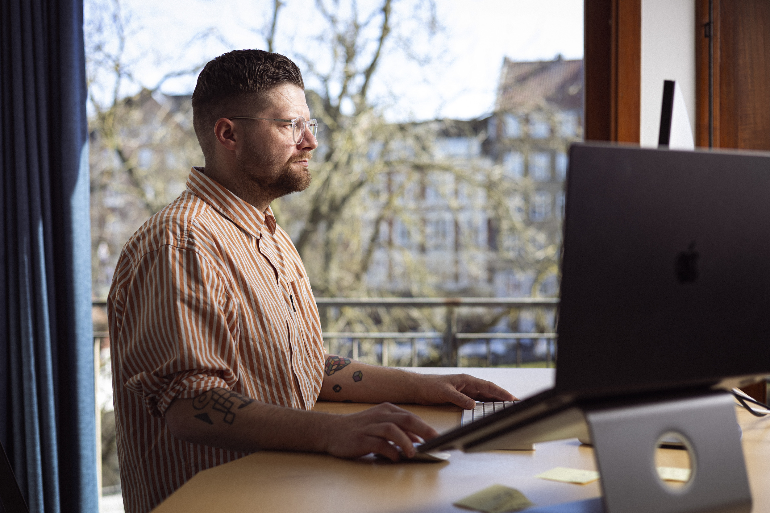 Mand arbejder på en computer ved et skrivebord med en balkonudsigt, iført en stribet skjorte.