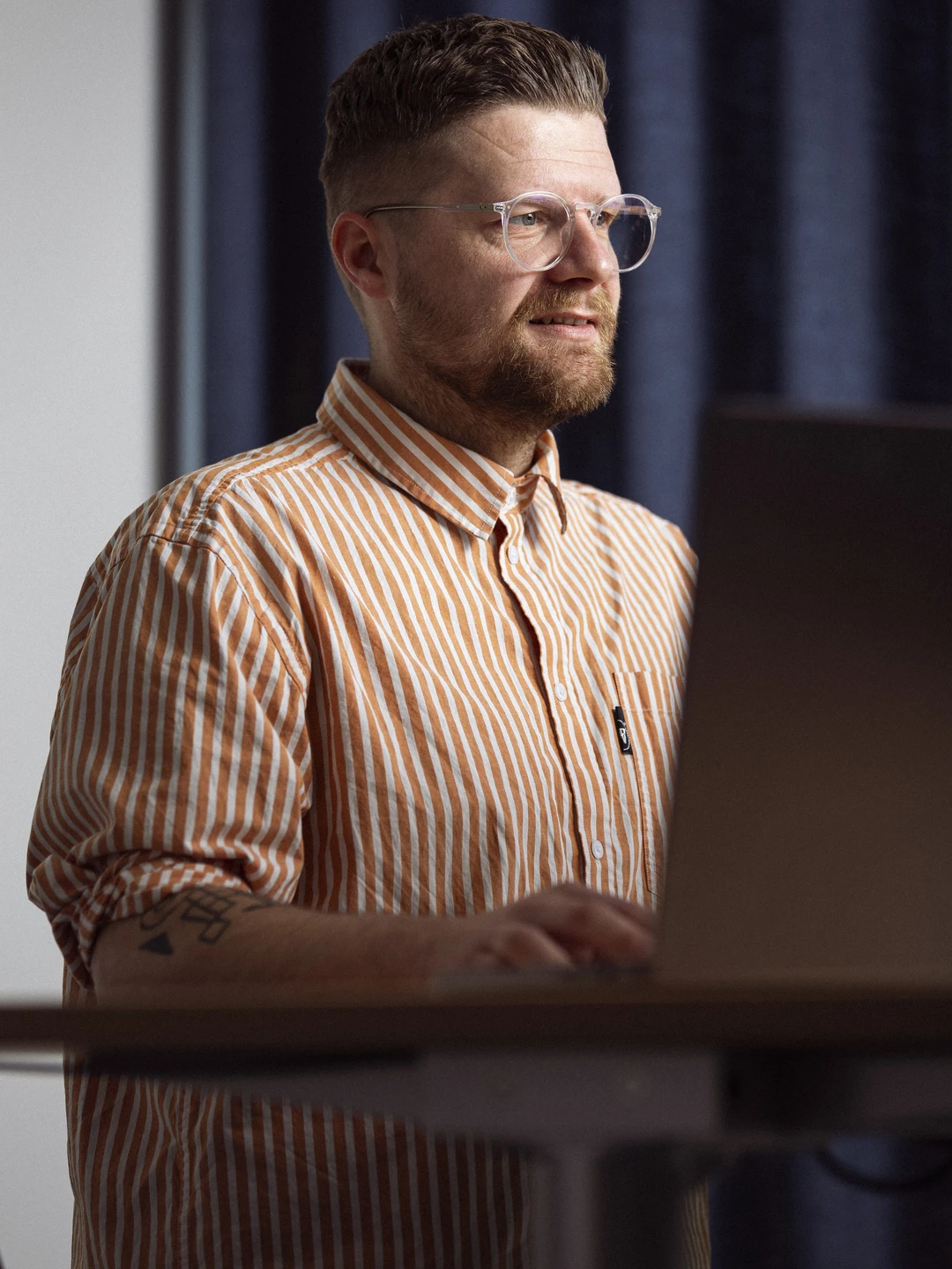 Mand arbejder på en laptop ved et stående skrivebord, iført en stribet orange og hvid skjorte, med briller og kort hår, i et rum oplyst af naturligt lys.