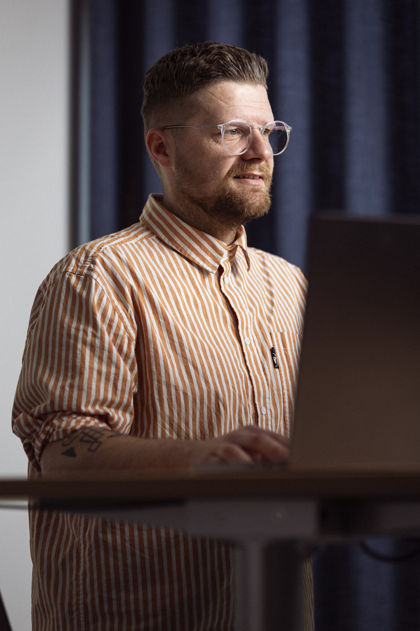 Mand arbejder på en laptop ved et stående skrivebord, iført en stribet orange og hvid skjorte, med briller og kort hår, i et rum oplyst af naturligt lys.