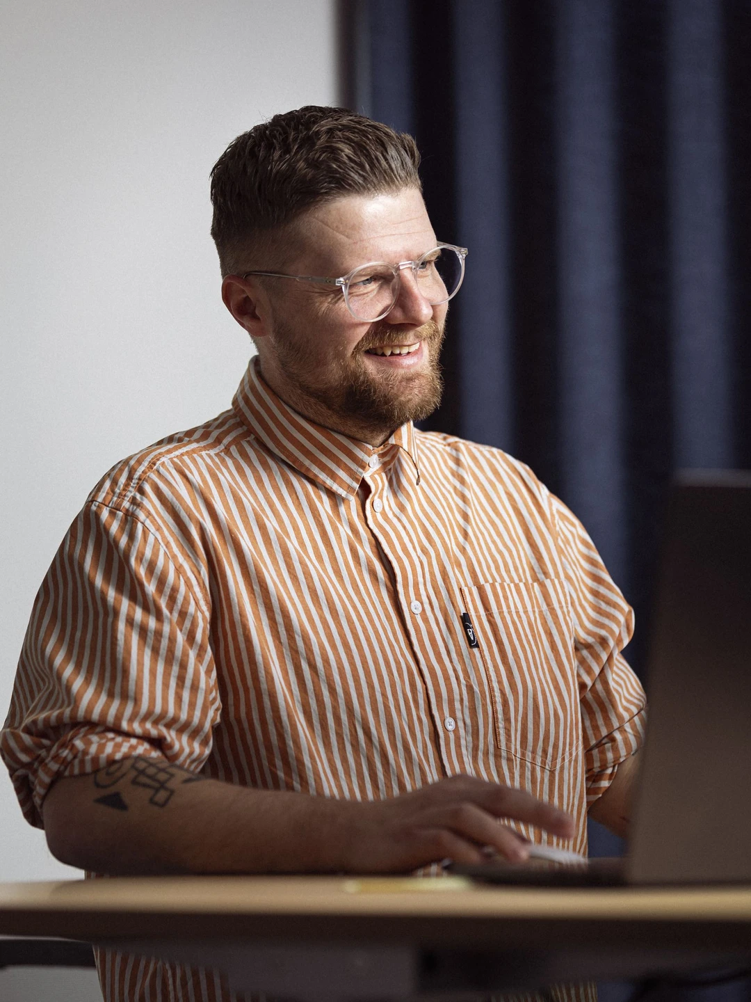Mand med briller og skæg smiler, mens han arbejder ved en computer, iført en stribet skjorte i orange og hvid, i et moderne kontormiljø.