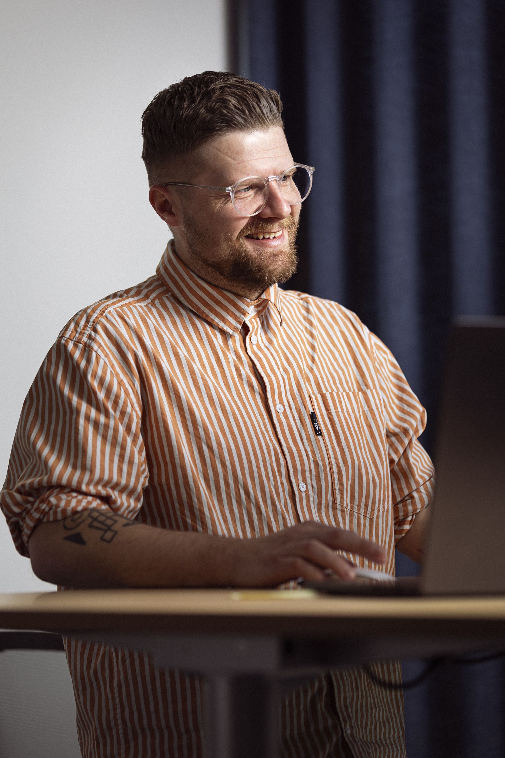 Mand med briller og skæg smiler, mens han arbejder ved en computer, iført en stribet skjorte i orange og hvid, i et moderne kontormiljø.