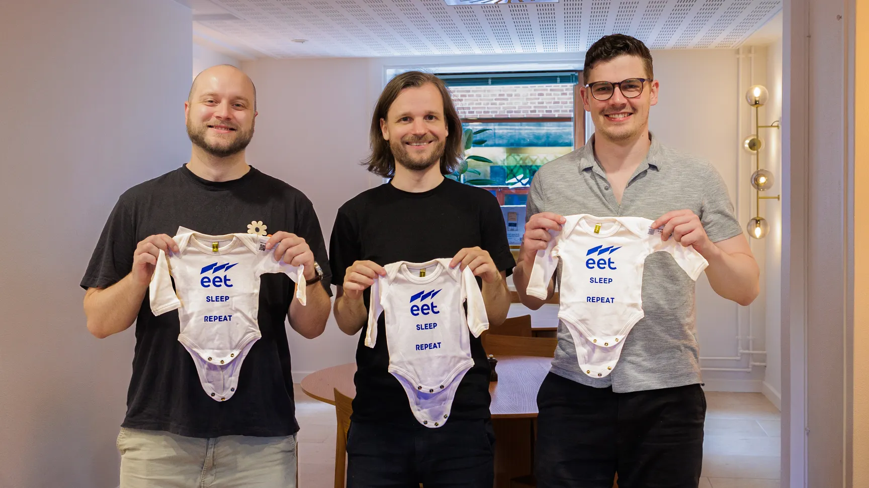 Tre mænd står i et lyst rum og holder op med hvidt babytøj med teksten "SLEEP REPEAT" og "eet". Rummet har et moderne design med en spisebord i baggrunden.
