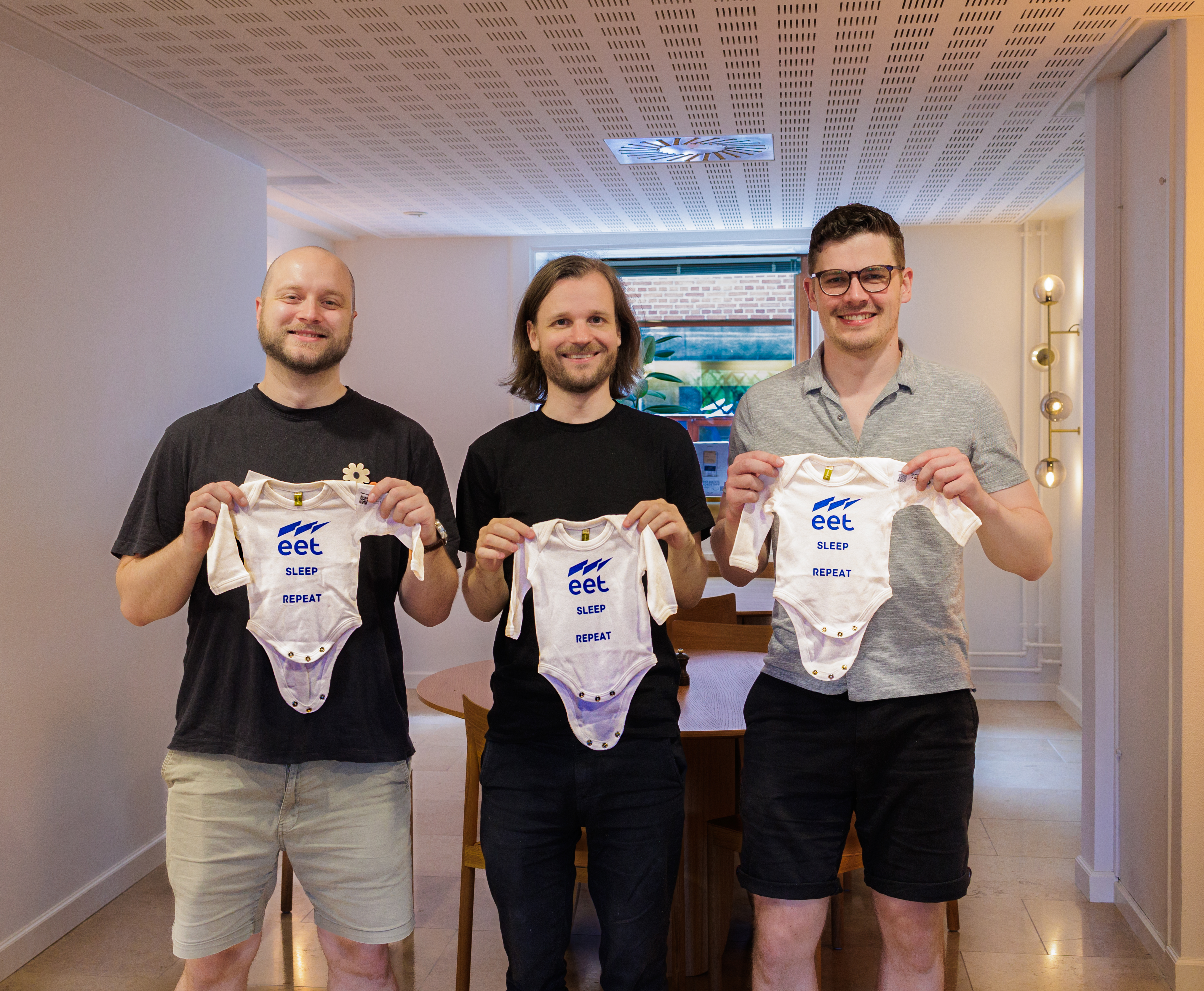Tre mænd står i et lyst rum og holder op med hvidt babytøj med teksten "SLEEP REPEAT" og "eet". Rummet har et moderne design med en spisebord i baggrunden.