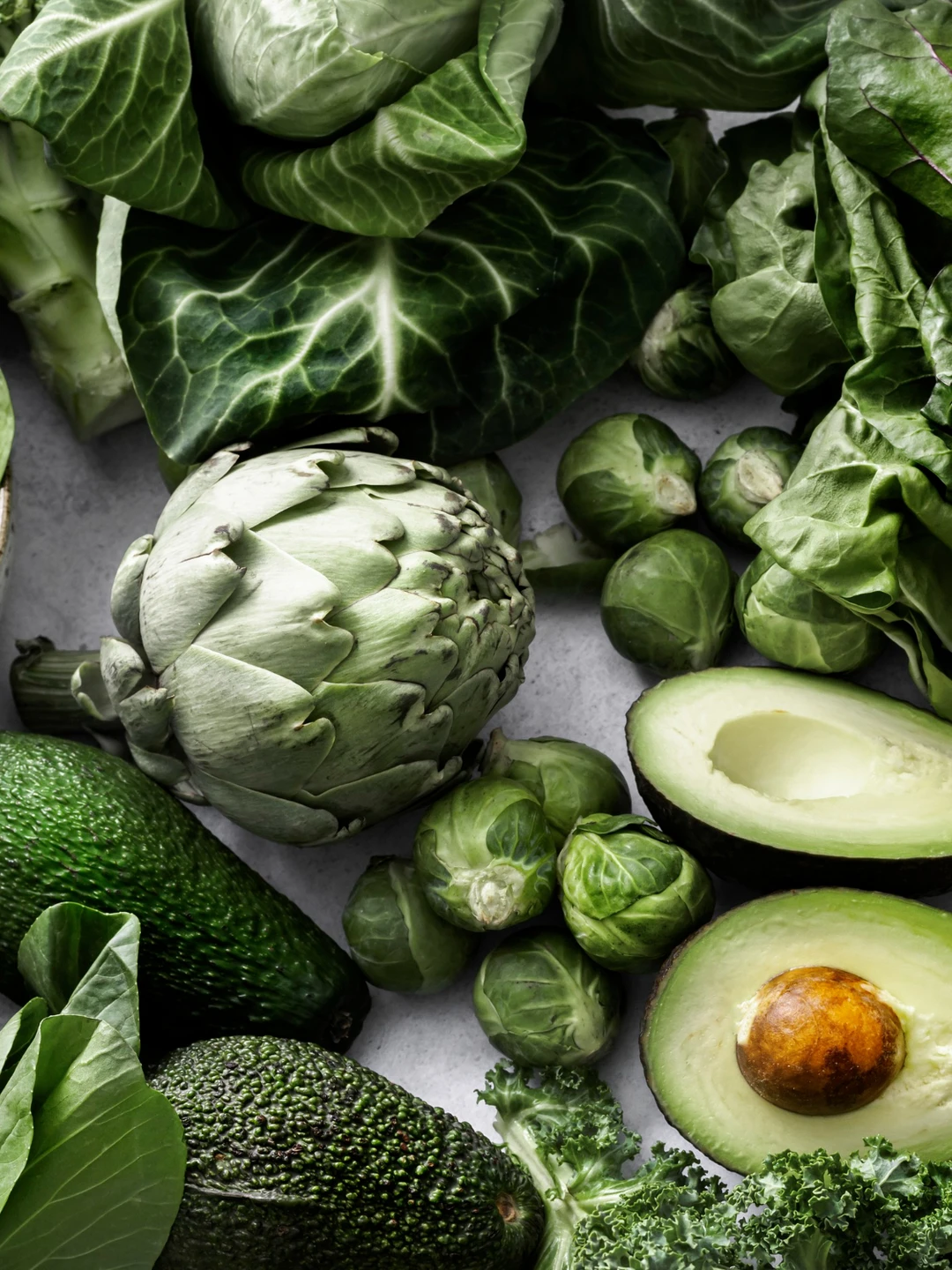En samling af friske, grønne grøntsager, herunder artiskokker, avocado, broccoli, rosenkål og forskellige typer bladgrønt på en lys baggrund.