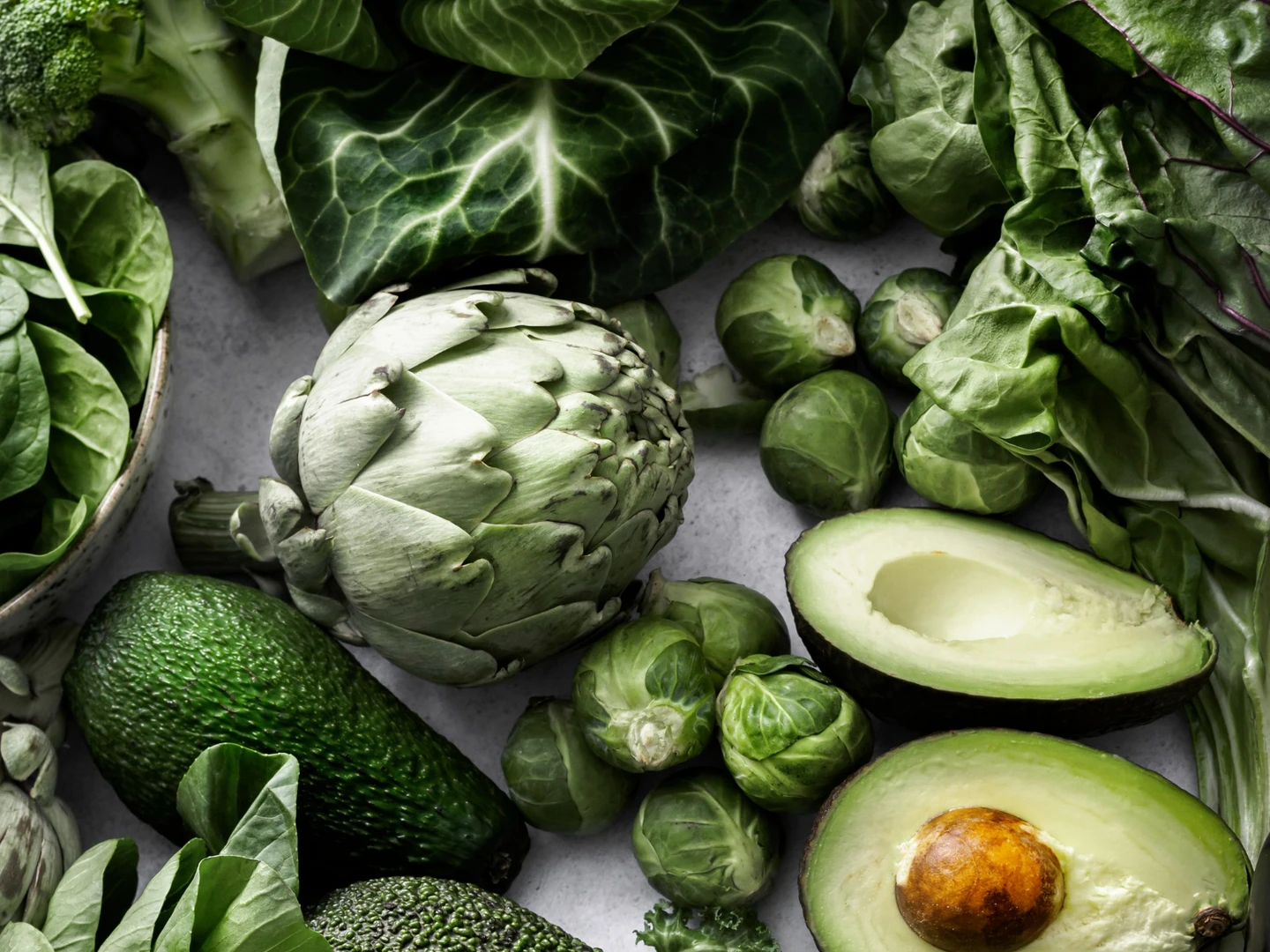 En samling af friske, grønne grøntsager, herunder artiskokker, avocado, broccoli, rosenkål og forskellige typer bladgrønt på en lys baggrund.