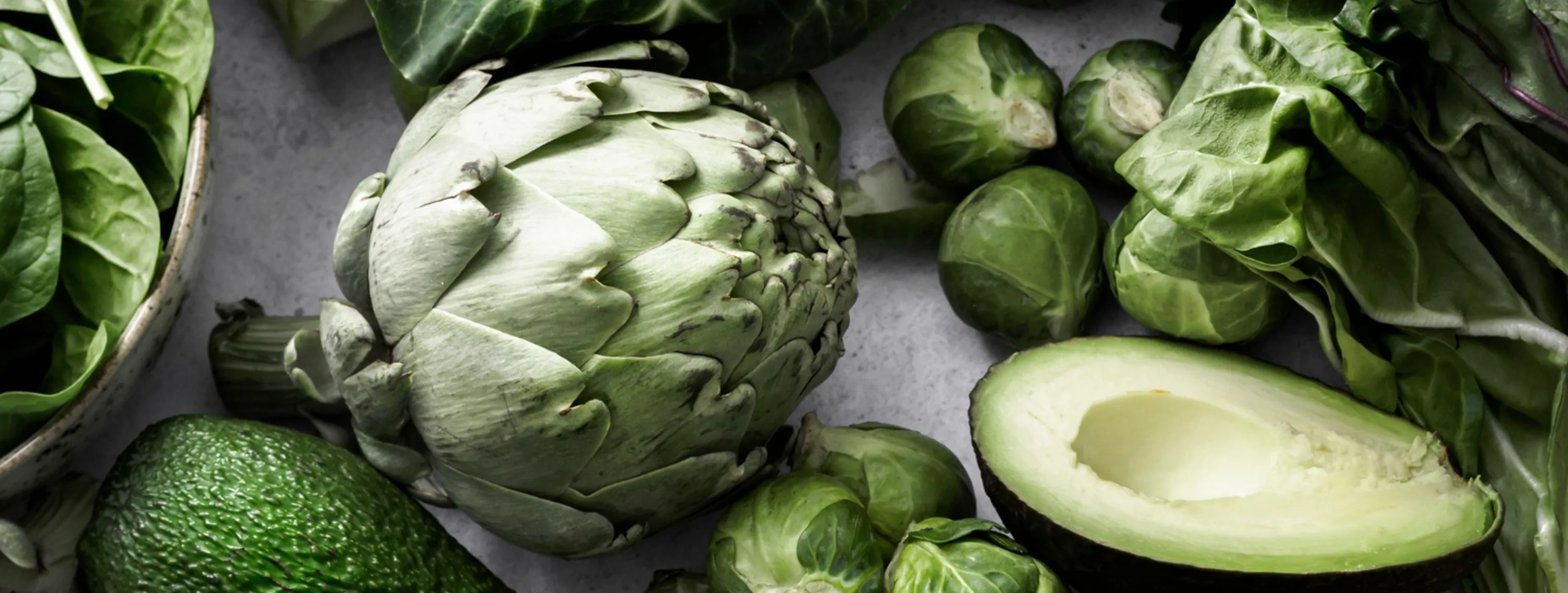 En samling af friske, grønne grøntsager, herunder artiskokker, avocado, broccoli, rosenkål og forskellige typer bladgrønt på en lys baggrund.