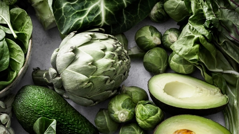 En samling af friske, grønne grøntsager, herunder artiskokker, avocado, broccoli, rosenkål og forskellige typer bladgrønt på en lys baggrund.
