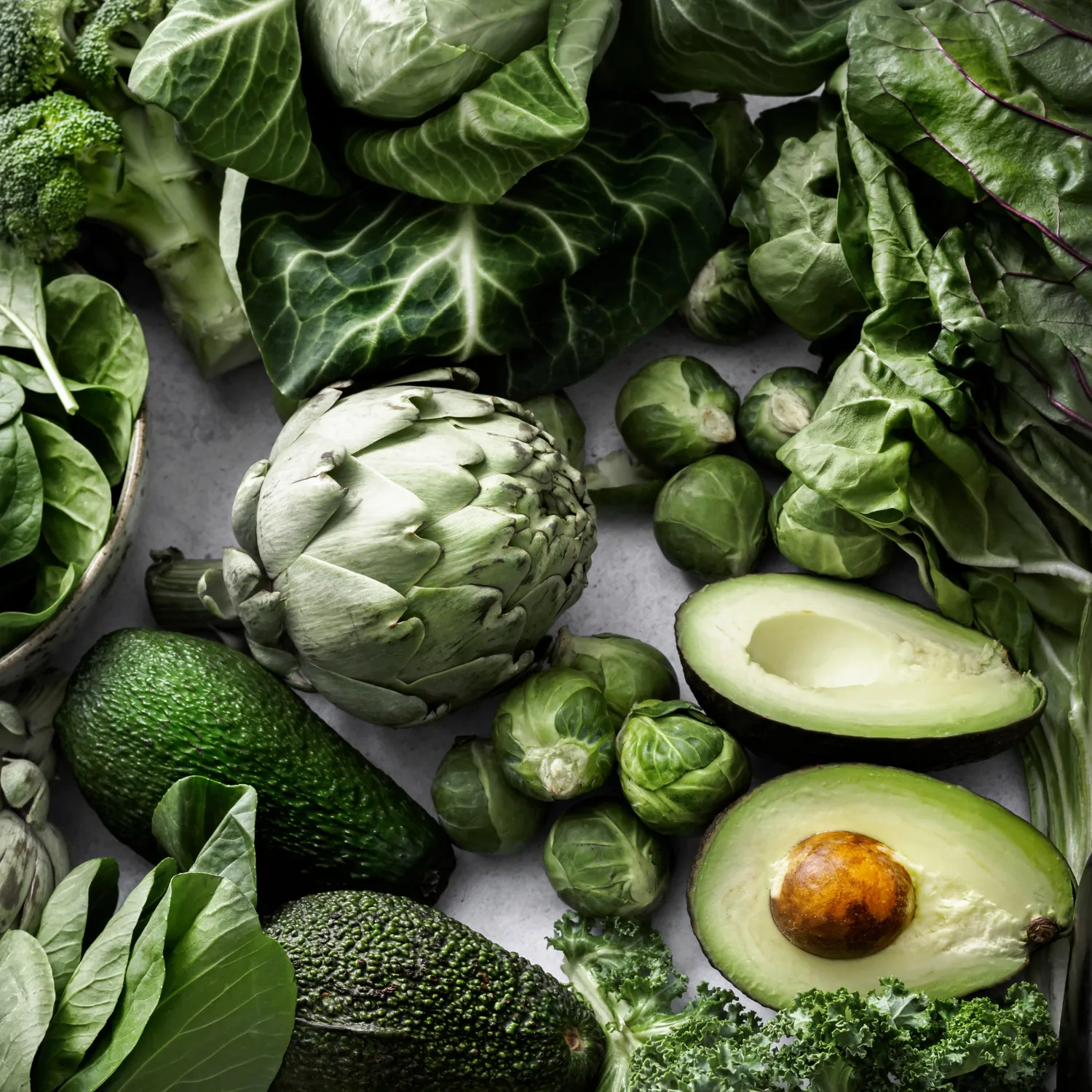 En samling af friske, grønne grøntsager, herunder artiskokker, avocado, broccoli, rosenkål og forskellige typer bladgrønt på en lys baggrund.