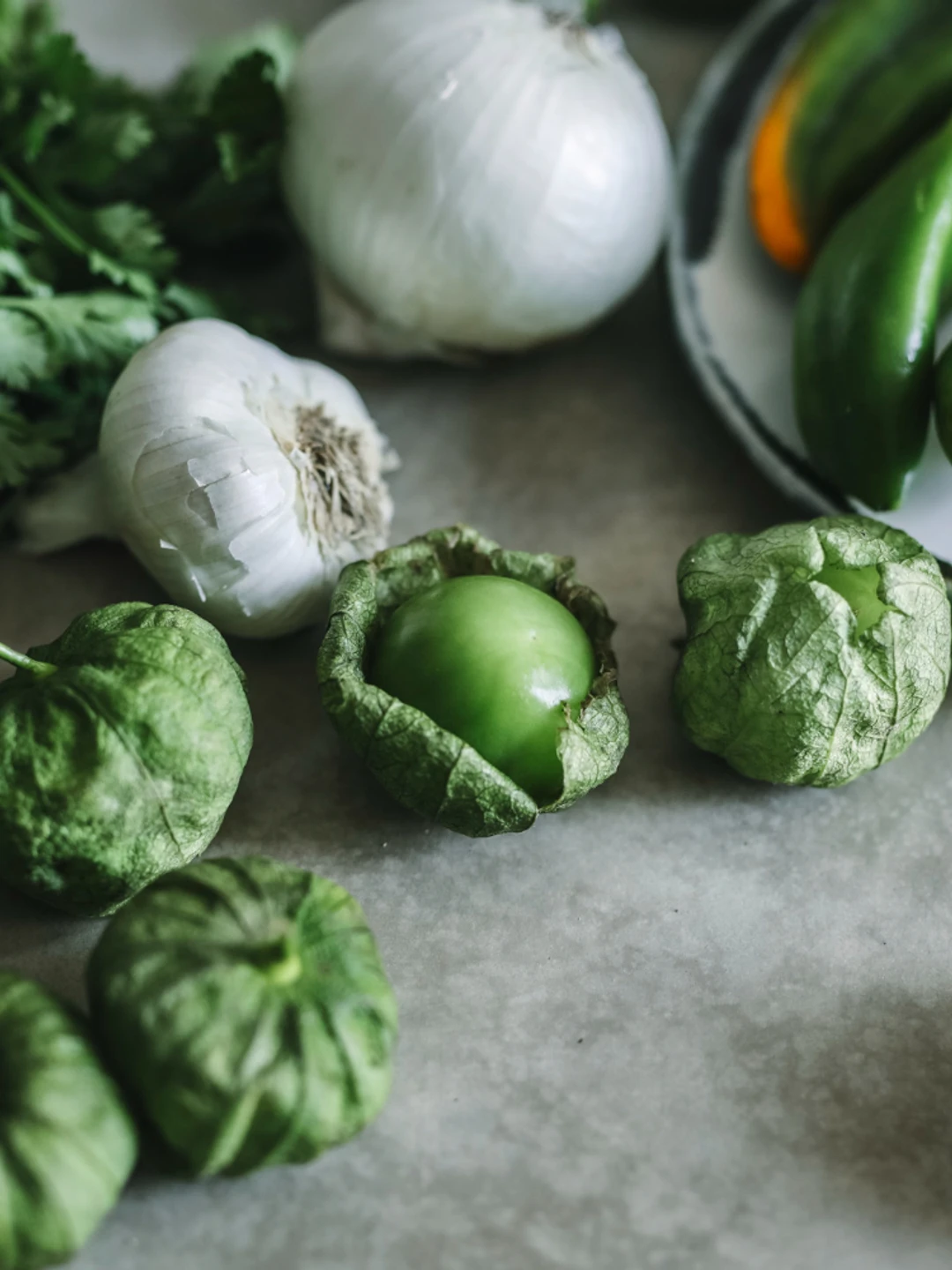 Friske grønne grøntsager herunder koriander, hvidløg, tomatillos og grønne chili på en bordplade.