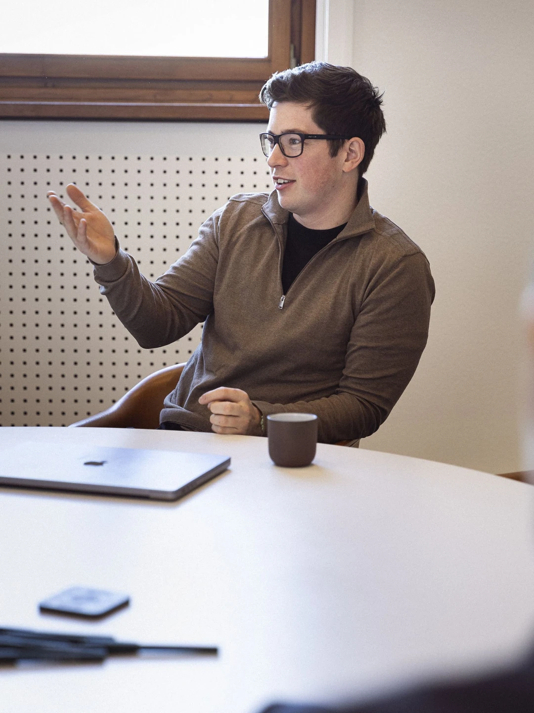 Ung mand med briller, iført en grå sweater, sidder ved et mødebord og gestikulerer mens han taler. En bærbar computer, en mobiltelefon og et krus med kaffe er synlige på bordet.
