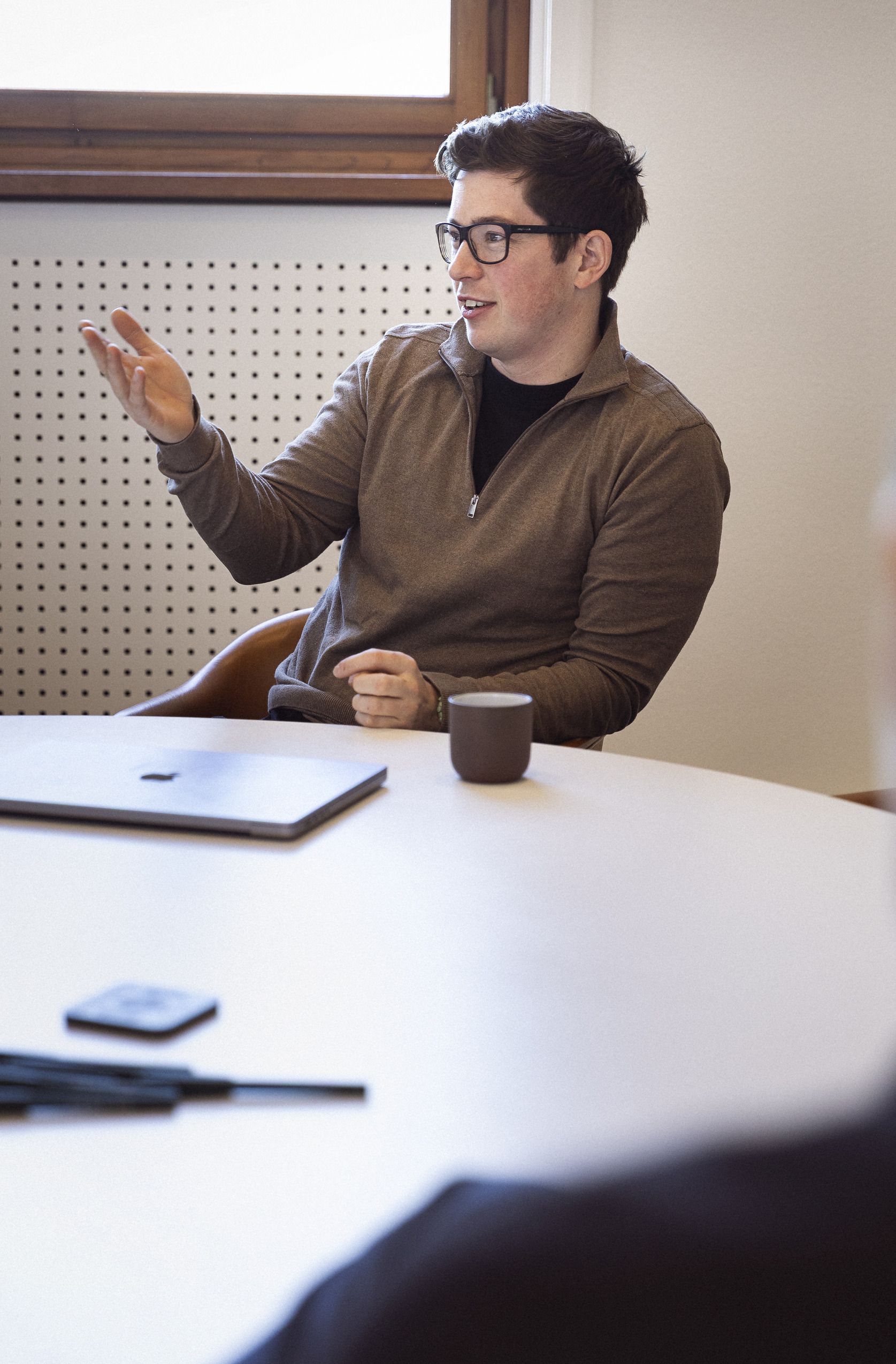 Ung mand med briller, iført en grå sweater, sidder ved et mødebord og gestikulerer mens han taler. En bærbar computer, en mobiltelefon og et krus med kaffe er synlige på bordet.