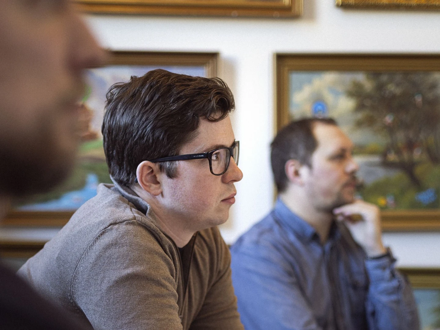 A group of individuals engaged in a focused discussion or meeting, with one person in the foreground visible from the side, wearing glasses and a brown sweater, while two others are partially visible in the background. The setting features framed artworks on the wall.