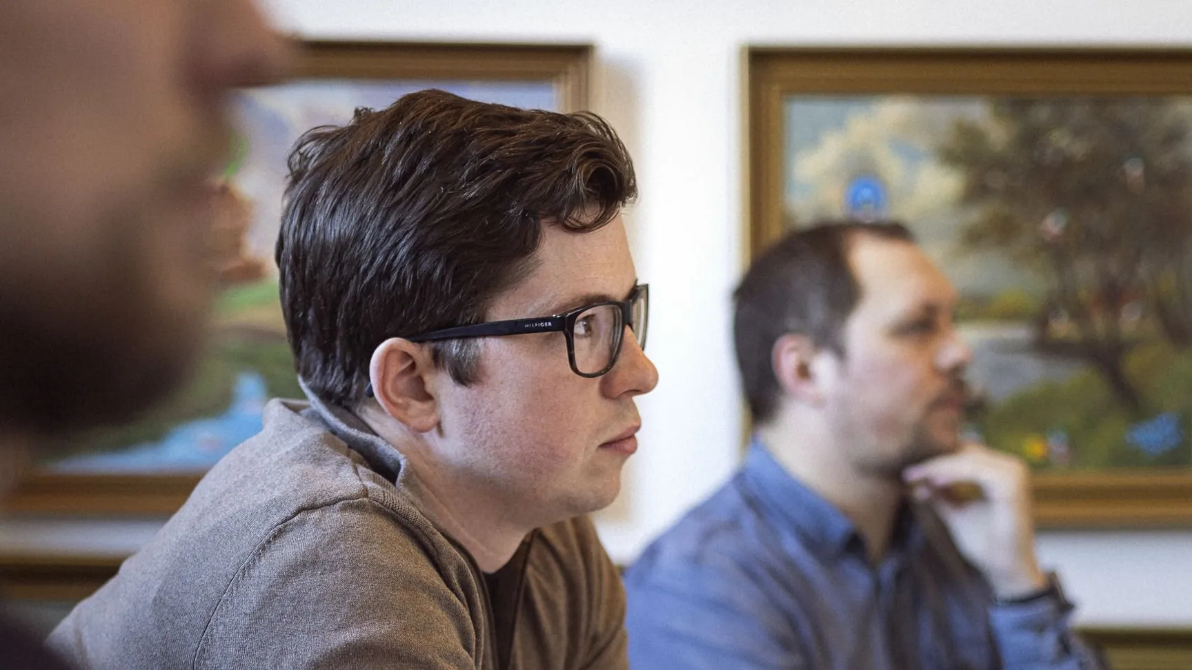 Tre mænd sidder ved et bord og lytter opmærksomt, mens de diskuterer. I baggrunden ses malerier på væggen, og der er fokus på mændenes ansigtsudtryk og hænder, der viser koncentration.